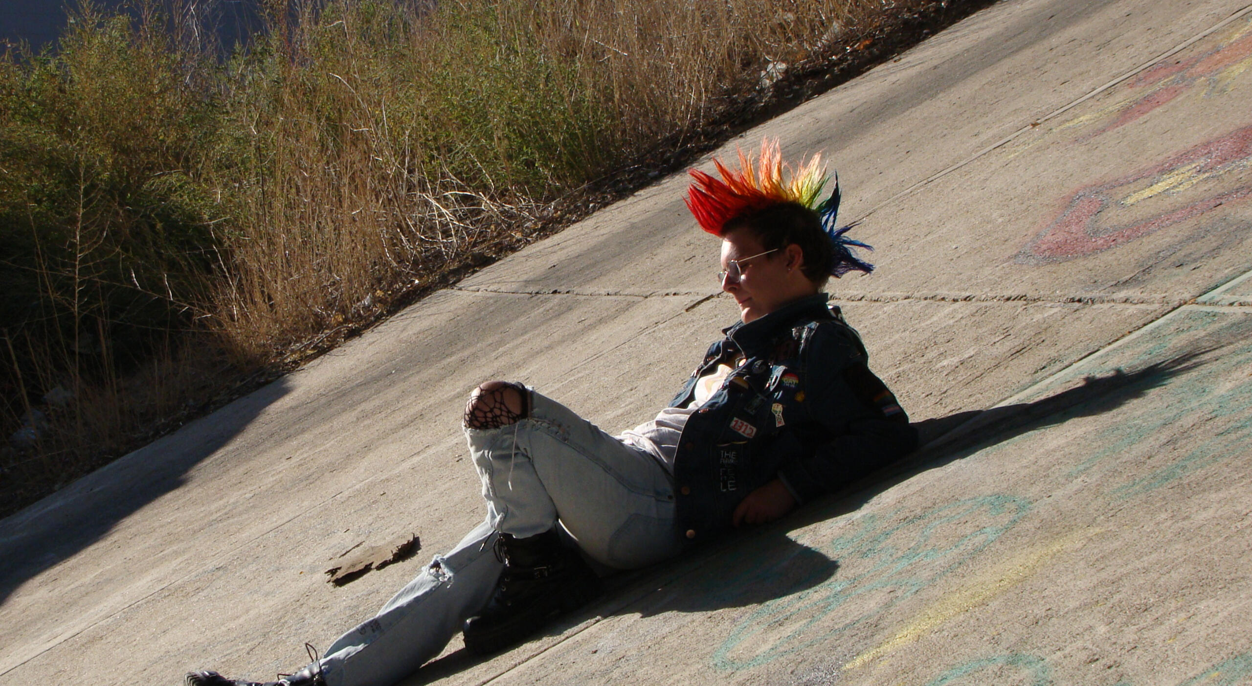 A photo of a young man with a rainbow, spiked mohawk lounging on a concrete slope. He is wearing a battle jacket with various pins and patches, as well as ripped jeans, fishnet tights, boots, and glasses. There are intense shadows being cast on him, and a